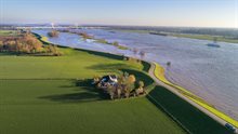 Dronefoto van rivierlandschap met hoogwater, dijk, boerderij en brug.